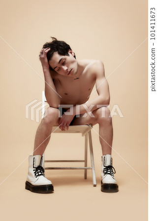 Thoughtful look. Young man sitting on chair shirtless in boots, holding head with hand, posing against studio background 101411673