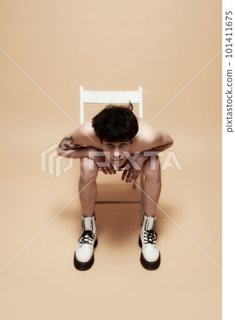 Young asian guy in underwear and stylish boots, posing shirtless on chair against light brown studio background 101411675