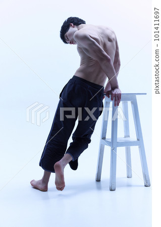 Young man with relief, beautiful body, posing shirtless in black trousers on high chair against white studio background. Back view 101411697