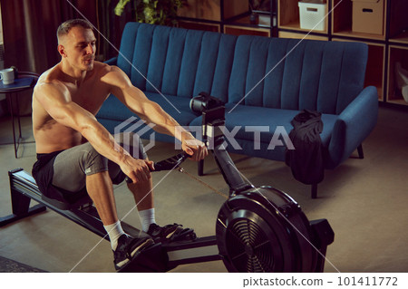 Strong, sportive, shirtless man with muscular body doing exercises with stationary rowing machine. Home gym Strong, sportive, shirtless man with muscular body doing exercises with stationary rowing machine. Home gym 101411772