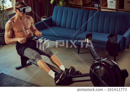 Strong young man with relief, muscular, fit, shirtless body training at home stationary rowing machine, in VR glasses. Modern style Strong young man with relief, muscular, fit, shirtless body training at home stationary rowing machine, in VR glasses. Modern style 101411773