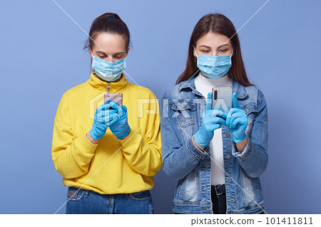 Indoor shot of attentive concentrated two women holding their smartphones, reading news about coronavirus, wearing antibacterial masks and gloves, being in quarantine. Self-isolation concept. 101411811