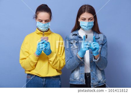 Horizontal indoor shot of beautiful curious young friends standing isolated over blue background in studio, using smartphones, being attentive, wearing masks and gloves. Coronavirus outbreak concept. 101411812