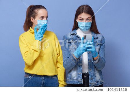 Portrait of two shocked emotional women wearing casual clothes, antibacterial masks and gloves, protecting themselves from coronavirus, using smartphone, checking news about virus. Covid 19 concept. Portrait of two shocked emotional women wearing casual clothes, antibacterial masks and gloves, protecting themselves from coronavirus, using smartphone, checking news about virus. Covid 19 concept. 101411816