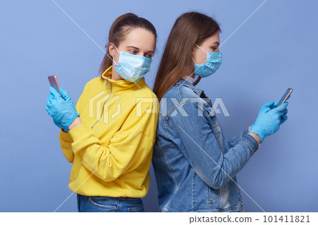 Picture of two busy curious attractive females standing back to back holding their smartphones, using devices, reading news, wearing antibacterial masks and gloves. People and quarantine concept. Picture of two busy curious attractive females standing back to back holding their smartphones, using devices, reading news, wearing antibacterial masks and gloves. People and quarantine concept. 101411821