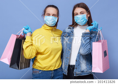 Indoor portrait of pleasant good looking lovely females looking directly at camera, hugging each other, spending time together while shopping, taking prevention measures, being in masks and gloves. 101411826