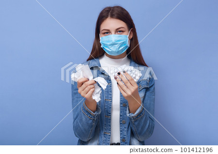 Indoor shot of sick lady wearing casual clothing and protective mask, posing isolated over blue studio wall, holding pills in hands, looking directly at camera. Corona virus, health care concept. Indoor shot of sick lady wearing casual clothing and protective mask, posing isolated over blue studio wall, holding pills in hands, looking directly at camera. Corona virus, health care concept. 101412196