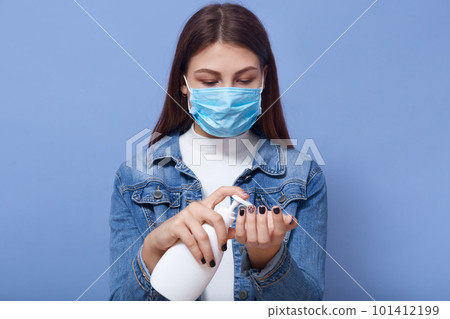 Image of careful young female standing isolated over blue background in studio, holding bottle with liquid, using antiseptic to damage bacterias, hygiene measures, in quarantine. Covid 19 concept. 101412199