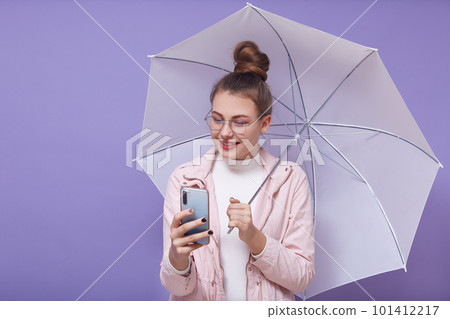 Portrait of good looking beautiful young girl holding umbrella and smartphone, wearing accessories, making selfie, shooting video, using social networking sites, having bunch on head. Youth concept. 101412217
