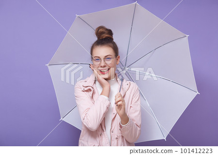 Close up portrait of beautiful young girl with hair bun, wearing white shirt, pink jacket and eyewear, posing with white umbrella in studio, cute female with charming smile looking at camera. 101412223