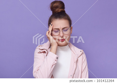 Indoor shot of young woman having headache, putting hand on temple, keeping eyes closed, standing isolated over lilac background, girl wearing casual clothing. Concept of problems, fever, health care. Indoor shot of young woman having headache, putting hand on temple, keeping eyes closed, standing isolated over lilac background, girl wearing casual clothing. Concept of problems, fever, health care. 101412264