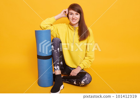 Shot of fitness woman sitting with exercise mat and looking directly at camera, adorable female working out at fitness club, wearing stylish yellow hoody and black leggins. Fitness and sport concept. Shot of fitness woman sitting with exercise mat and looking directly at camera, adorable female working out at fitness club, wearing stylish yellow hoody and black leggins. Fitness and sport concept. 101412319
