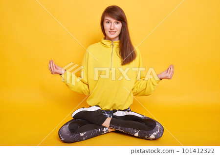 Indoor shot of slim sportswoman sitting on floor in lotus pose, doing yoga, female wearing leggins and hoody, attractive teenager posing isolated over yellow background. Fitness and sport concept. 101412322