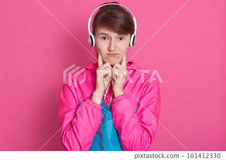 Studio shot of young cheerful Caucasian sporty girl in sportwear with white headphone on head, listening to music, looks upset, keeps fingers on cheek. Healthy lifestyle, fitness, good health concept. 101412330