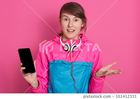 Impressed shocked dark haired hipster girl surprised to listen shocking news, has headphones around neck and smart phone wirh blank screen in hand, isolated over pink studio wall. Copy space. 101412331