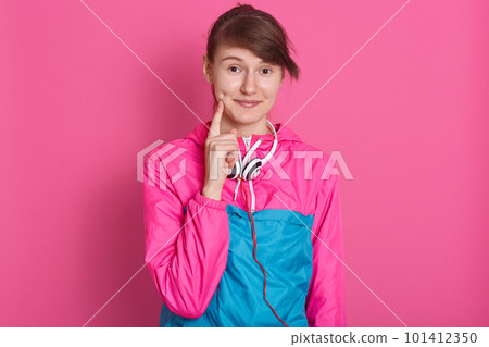 Close up portrait of young woman standing smiling and keeping fingers on cheeks while posing against yellow studio background, lady wearing blue and pink sportwear, looking directly at camera. Close up portrait of young woman standing smiling and keeping fingers on cheeks while posing against yellow studio background, lady wearing blue and pink sportwear, looking directly at camera. 101412350