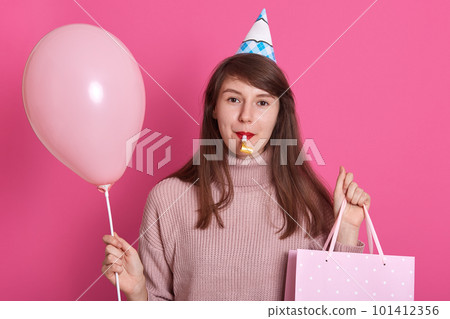 Portrait of beautiful young woman with party whistle and birthday hat, dresses pink sweater, model posing over pastel pink background, holding air baloons. Birthday, holiday party, people concept. Portrait of beautiful young woman with party whistle and birthday hat, dresses pink sweater, model posing over pastel pink background, holding air baloons. Birthday, holiday party, people concept. 101412356