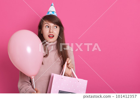 Happy brunette girl with pink balloon and bithday present in hands, keeps mouth opened, being in surprise, looks aside with astinished facial expression, copy space for advertisment or promotion. Happy brunette girl with pink balloon and bithday present in hands, keeps mouth opened, being in surprise, looks aside with astinished facial expression, copy space for advertisment or promotion. 101412357