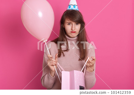 Closeup portrait of beautiful disappointed girl with gift package and balloon, wearing pink sweater and birthday hat, posing isolated over rosy background with pouty lips, having unhappy expression. Closeup portrait of beautiful disappointed girl with gift package and balloon, wearing pink sweater and birthday hat, posing isolated over rosy background with pouty lips, having unhappy expression. 101412359