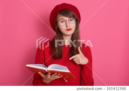 Horizontal shot of woman with orange note book pointing at its pages with her index finger. Attractive female wearing red sweater, beret and glasses, having perfect skin and bright pomade makeup. Horizontal shot of woman with orange note book pointing at its pages with her index finger. Attractive female wearing red sweater, beret and glasses, having perfect skin and bright pomade makeup. 101412390