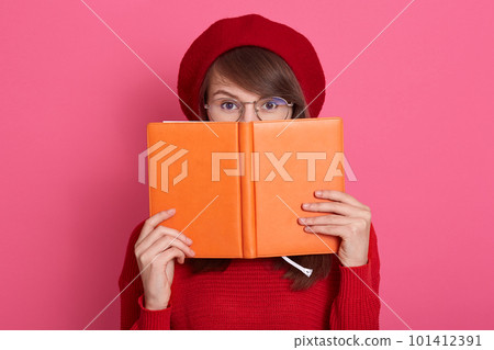 Close up portrait of young woman hides her face behind her notebook orange notebook, charming female wears red sweater, beret and spectacles, looks scared behind her organaizer, isolated on pink wall. Close up portrait of young woman hides her face behind her notebook orange notebook, charming female wears red sweater, beret and spectacles, looks scared behind her organaizer, isolated on pink wall. 101412391