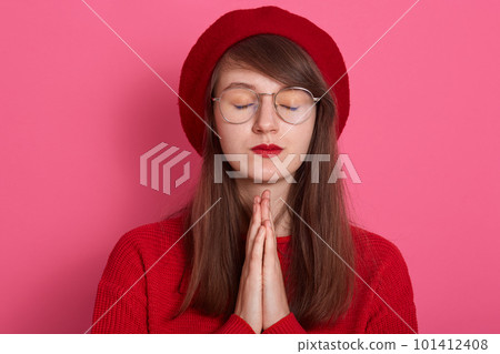 Image of beautiful woman wearing red clothing and beret, keeps palms pressed together in front of her, pouts lips, has regretful expression, keeping eyes closed, isolated over pink studio background. Image of beautiful woman wearing red clothing and beret, keeps palms pressed together in front of her, pouts lips, has regretful expression, keeping eyes closed, isolated over pink studio background. 101412408
