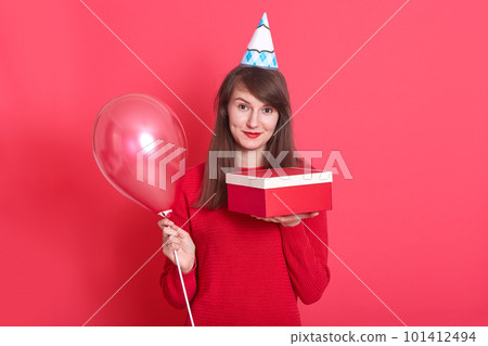 Indoor picture of magnetic lovely young woman having holiday cap on head, holding red baloon and red and white present box, looking directly at camera, wearing bright red sweater. Holiday concept. Indoor picture of magnetic lovely young woman having holiday cap on head, holding red baloon and red and white present box, looking directly at camera, wearing bright red sweater. Holiday concept. 101412494
