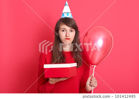 Studio shot of young woman girl posing isolated over red background. Valentine's Day, birthday concept, female holding air balloons and present box, looks upset and sad, lady wears beret and sweater. Studio shot of young woman girl posing isolated over red background. Valentine's Day, birthday concept, female holding air balloons and present box, looks upset and sad, lady wears beret and sweater. 101412495
