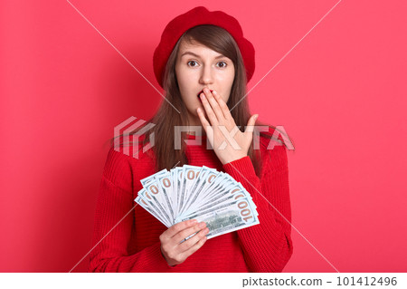 Photo of rich beautiful woman dresses red sweater and beret holding fan of dollar money isolated over background, looks astonished, covering his mouth with palm, attractive female posing in studio. Photo of rich beautiful woman dresses red sweater and beret holding fan of dollar money isolated over background, looks astonished, covering his mouth with palm, attractive female posing in studio. 101412496
