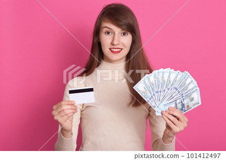 Closeup portrait of dark haired woman holding credit card and fan of money, posing isolated over pink studio background, lady looking directly at camera, looks happy, girl doing shopping online. Closeup portrait of dark haired woman holding credit card and fan of money, posing isolated over pink studio background, lady looking directly at camera, looks happy, girl doing shopping online. 101412497