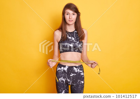 Photo of slim lady wearing sports clothing, holding measure tape, measuring her wrist, comparing her results after fitness exercises, posing against yellow studio wall. Sport and keeping fit concept. 101412517
