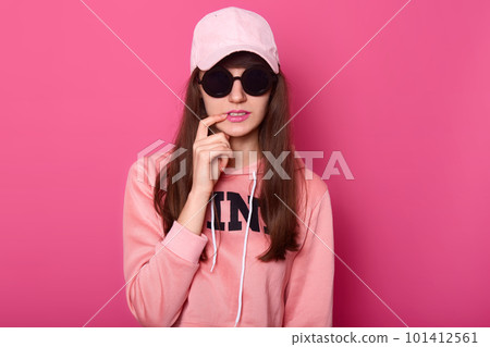 Young dark haired teenager girl wearing stylish pink hoodie, cap and black subglasses, touches her lip with fore finger,flirts with boyfriend. Studio portrait of fashinable female. People concept. 101412561