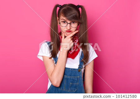 Childish charismatic young lady puts her hand on face, thinking over her plans, makes frowny face. Funny girl with bangs and long pigtails stands straight isolated over bright pink background. Childish charismatic young lady puts her hand on face, thinking over her plans, makes frowny face. Funny girl with bangs and long pigtails stands straight isolated over bright pink background. 101412580
