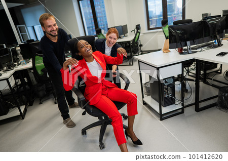 Caucasian red-haired woman, bearded caucasian man rolled African American young woman on office chair. Colleagues have fun at work. Caucasian red-haired woman, bearded caucasian man rolled African American young woman on office chair. Colleagues have fun at work. 101412620