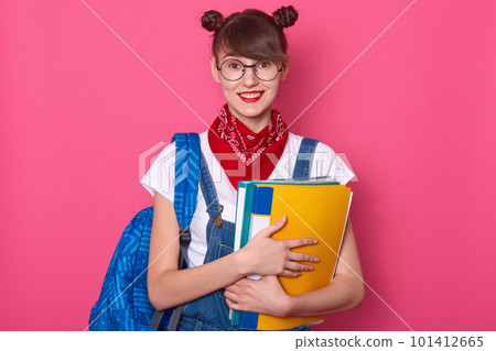 Horizontal shot of peasant looking woman with two hair buns, has delighted expression, holding blue and yellow folder, dresses casual clothing, posing after having classes, isolated over pink wall. Horizontal shot of peasant looking woman with two hair buns, has delighted expression, holding blue and yellow folder, dresses casual clothing, posing after having classes, isolated over pink wall. 101412665