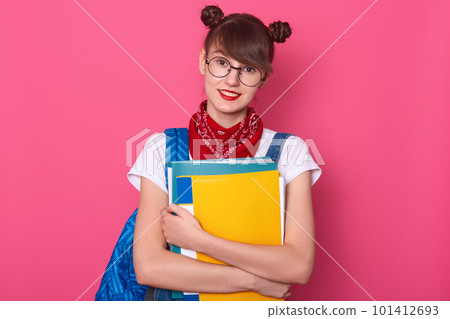 Horizontal shot of smiling lady student wears eyeglasses, red bandana over her neck, white casual t shirt, stands with folder in hands, happily looks directly at camera, poses on pink background. Horizontal shot of smiling lady student wears eyeglasses, red bandana over her neck, white casual t shirt, stands with folder in hands, happily looks directly at camera, poses on pink background. 101412693