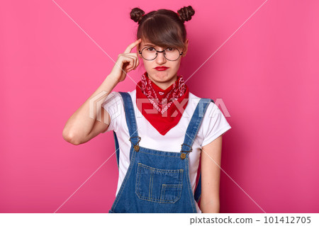 Close up portrait of brunette student, wears round eyewear and red bandana on neck, curves lips, keeps fore finger on temple, poses against pink background. Copy space for your advertisment. 101412705