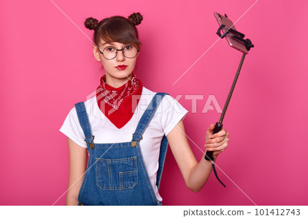 Image of beautiful girl in white t-shirt and denim overals, takes a selfie with her smart phone, uses selfie stick, isolated over rose background. Copy space for promotion text. Technology concept. 101412743