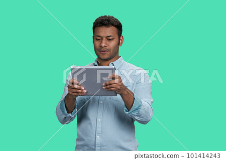 Portrait of a young thoughtful indian guy holding tablet pc. South asian man readin gsomething on his device. Isolated on green background. Portrait of a young thoughtful indian guy holding tablet pc. South asian man readin gsomething on his device. Isolated on green background. 101414243