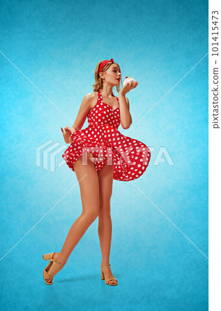 Sweet tooth. Full-length portrait of young pretty blonde girl in stylish red dress posing against blue background, eating cake. Pin-up style 101415473