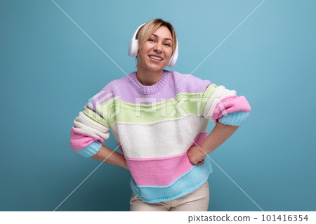 cute attractive blond woman in a striped sweater in big wireless headphones enjoys a podcast on a 101416354
