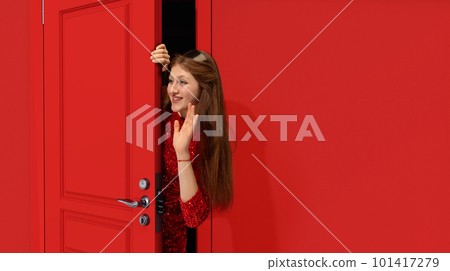 Stylish, beautiful teen girl in trendy red sparkling dress peeking out red door and waving to friends. Party, meeting, celebration Stylish, beautiful teen girl in trendy red sparkling dress peeking out red door and waving to friends. Party, meeting, celebration 101417279