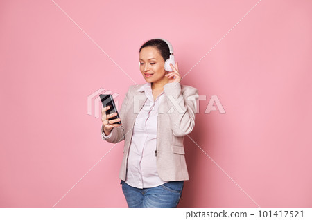 Attractive multi-ethnic middle-aged pregnant woman wearing wireless headphones, listening to music or watching newborn baby care video on the internet, isolated pink background. Maternity. Pregnancy Attractive multi-ethnic middle-aged pregnant woman wearing wireless headphones, listening to music or watching newborn baby care video on the internet, isolated pink background. Maternity. Pregnancy 101417521