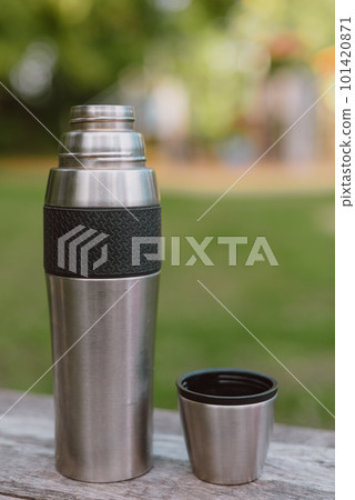 Stainless steel cup, mug with thermos stands on a wooden table in the forest on a green natural background. Thermos and mug with hot drink standing on wet wooden table after rain outdoors, close-up Stainless steel cup, mug with thermos stands on a wooden table in the forest on a green natural background. Thermos and mug with hot drink standing on wet wooden table after rain outdoors, close-up 101420871