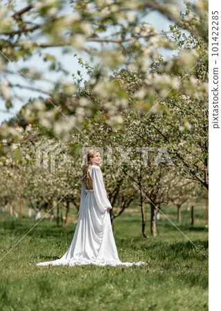 Blond blooming garden. A woman in a white dress walks through a blossoming cherry orchard. Long dress flies to the sides, 101422285