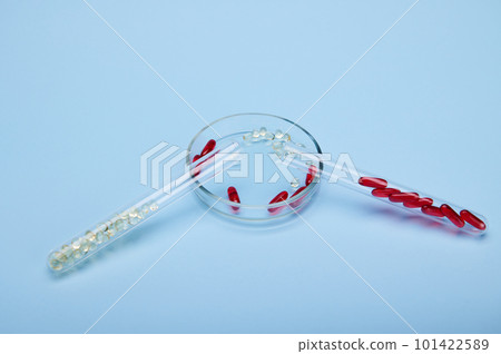 Soft translucent gelatin capsules of red and gold color with essential oils, omega-3, fat-soluble vitamins inside test tube and petri dish, isolated blue backdrop. Pharmaceutical industry. Still life Soft translucent gelatin capsules of red and gold color with essential oils, omega-3, fat-soluble vitamins inside test tube and petri dish, isolated blue backdrop. Pharmaceutical industry. Still life 101422589