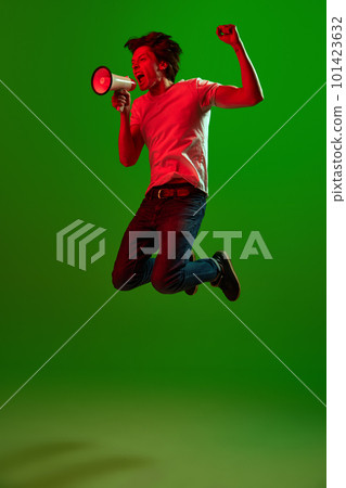 One young angry man, guy screaming through megaphone, jumping and threatens with a fist over green background in neon light. Breaking news 101423632