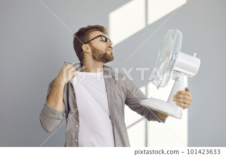 Young man suffers from summer heat and uses electric fan blowing fresh air in his face 101423633