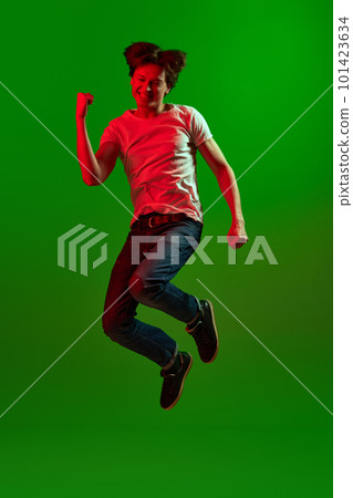 Studio shot of young guy, student screaming with exciting face and jumping over green background in neon light. Victory, he did it 101423634