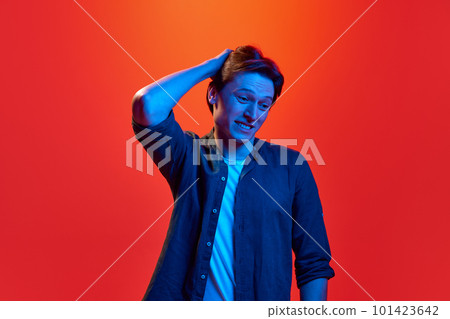 Portrait of young guy, man wearing shirt standing with uncertain facial expression over red-orange background in neon light. Doubtful guy 101423642